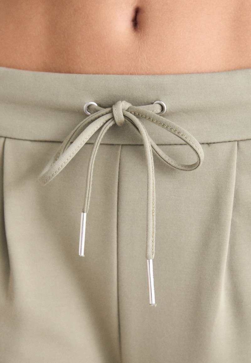 Hellgrüner Kordelzug mit silbernen Metallspitzen, zu einer Schleife gebunden, an einem weichen, strukturierten Bund mit Falten. Nahaufnahme des Stoffdetails.