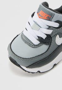 Nike sneaker, amely szürke szintetikus felsőrésszel rendelkezik fekete akcentusokkal, fehér fűzőkkel és egy kiemelkedő narancssárga logóval. Lekerekített orr, texturált részletekkel.