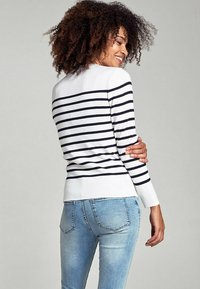Maglione bianco con righe orizzontali blu navy, maniche lunghe, polsini e orlo a coste. Indossato con jeans blu chiaro, che mostrano un look casual.