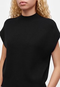 Femme aux cheveux blonds bouclés portant un pull noir sans manches à col montant, visible des épaules jusqu'au milieu du torse sur un fond uni.