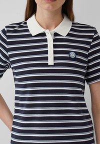 Marineblauwe en witte gestreepte polo met een witte kraag en een knoopsluiting met vier knopen. Heeft een ronde, blauwe logo op de linkerborst.