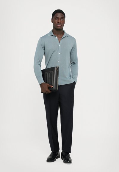 Homem que veste uma camisa azul clara com botões e calças escuras, segurando uma bolsa de couro preta contra um fundo branco.