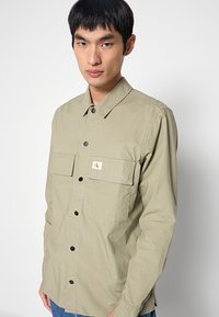 Hellgrünes Hemd mit Knopfleiste, zwei Fronttaschen, schwarzen Knöpfen und kleinem Logoaufnäher. Der Stoff hat eine strukturierte, legere Optik.