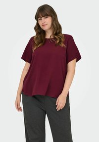 Mörkröd kortärmad topp med en avslappnad passform och rund halsringning, kombinerad med grå byxor. Slät tyg med en lätt struktur.