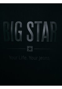 Melnas audums ar lieliem iespiestiem burtiem "BIG STAR" virs mazās zvaigznes ikonas un frāzi "Your Life. Your Jeans." mazākos iespiestos burtiņos.