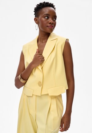 Kobieta z krótkimi naturalnymi włosami, ubrana w bezrękawnik w bladożółtym kolorze i dopasowane plisowane spodnie, ozdobiona kolczykami kołami i złotymi bransoletkami.