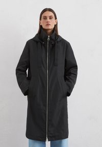 Parka noire à capuche avec fermeture éclair intégrale, deux poches avant et cordons de serrage réglables sur la capuche. Tissu lisse et non réfléchi.