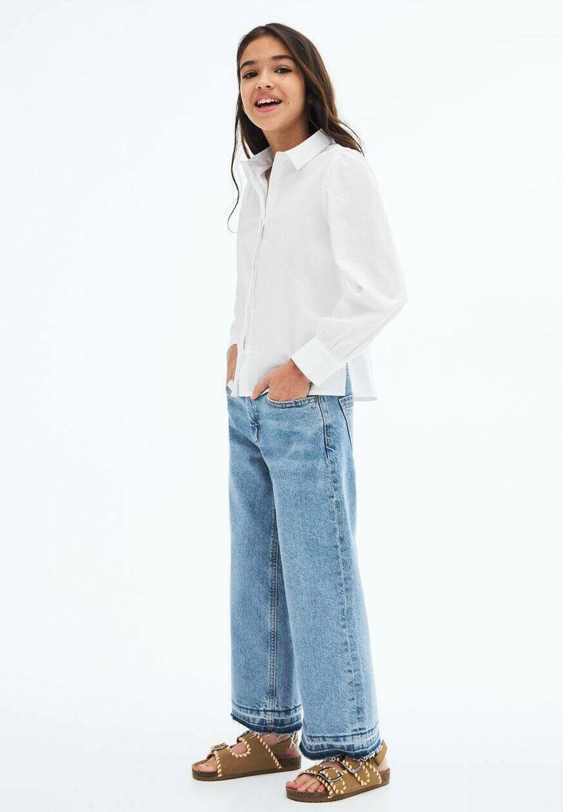 Valkoinen nappikauluspaita pitkillä hihoilla, vaaleansiniset leveälahkeiset farkut ja ruskeat sandaalit, joissa on hihnat ja niitit. Yksinkertainen muotoilu ja rento malli.