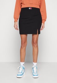 Svart ribbad minikjol med en sidoklyvning, bärs med en orange sweatshirt och ljusblå sneakers, vilket framhäver färgkontraster.