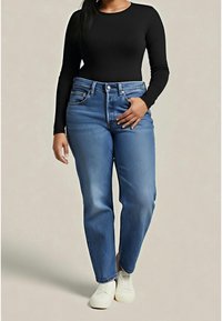 Femme portant un jean bleu, des baskets blanches et un haut noir à manches longues, debout avec une main dans la poche sur un fond uni.