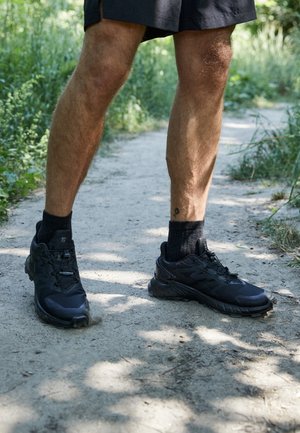 Zwarte sportieve schoenen met een gestructureerde bovenkant, gevoerde zool en veilige veters, gedragen met zwarte crew-sokken op een aarde pad omringd door groen.