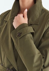 Trench-coat vert olive avec un col structuré, des poignets à boutons et une taille ceinturée, présentant un tissu doux et texturé.