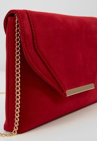 Pochette en daim rouge avec une bandoulière chaîne dorée, un design pliable et un accent métallique rectangulaire à l'avant. Texture lisse et finition douce.