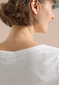 Biała bawełniana koszula z szerokim dekoltem i delikatną fakturą, podkreślająca obszar ramion i szyi. Widoczne krótkie, lekko kręcone włosy.
