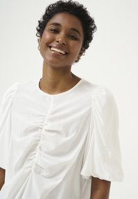 Blusa bianca con maniche a palloncino, caratterizzata da un design frontale arricciato. Tessuto leggero e liscio con una leggera texture. Stile minimalista, collo a giro.