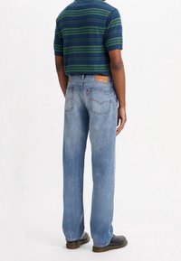 Man med ljusblå raka jeans, svarta skor och en marinblå polo med gröna ränder, stående vänd bort.