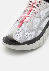 Scarpa sportiva bianca e grigia con lacci con accenti rossi, tomaia in mesh traspirante, punta nera e suola spessa e texturizzata.