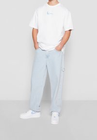 T-shirt bianco in cotone con testo ricamato blu, jeans in denim chiaro e larghi, e sneakers bianche con dettagli a scacchi blu.