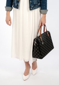 Black handbag with quilted pattern, brown handles, held by a person in a white dress and denim jacket, with white pointed heels.
