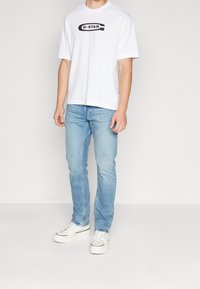 Vit bomull t-shirt med en svart "G-STAR" logotyp, i kombination med ljusblå denimjeans och vita sneakers med svarta detaljer.