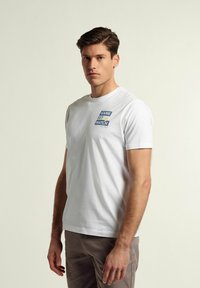 Camiseta blanca de algodón con texto gráfico azul y amarillo "GAME SET MATCH" en el frente, cuello redondo y mangas cortas.