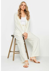Blazer chiaro in crema e pantaloni a gamba larga in una miscela di lino, abbinati a sandali bianchi con cinturini. Sgabello in legno sullo sfondo.