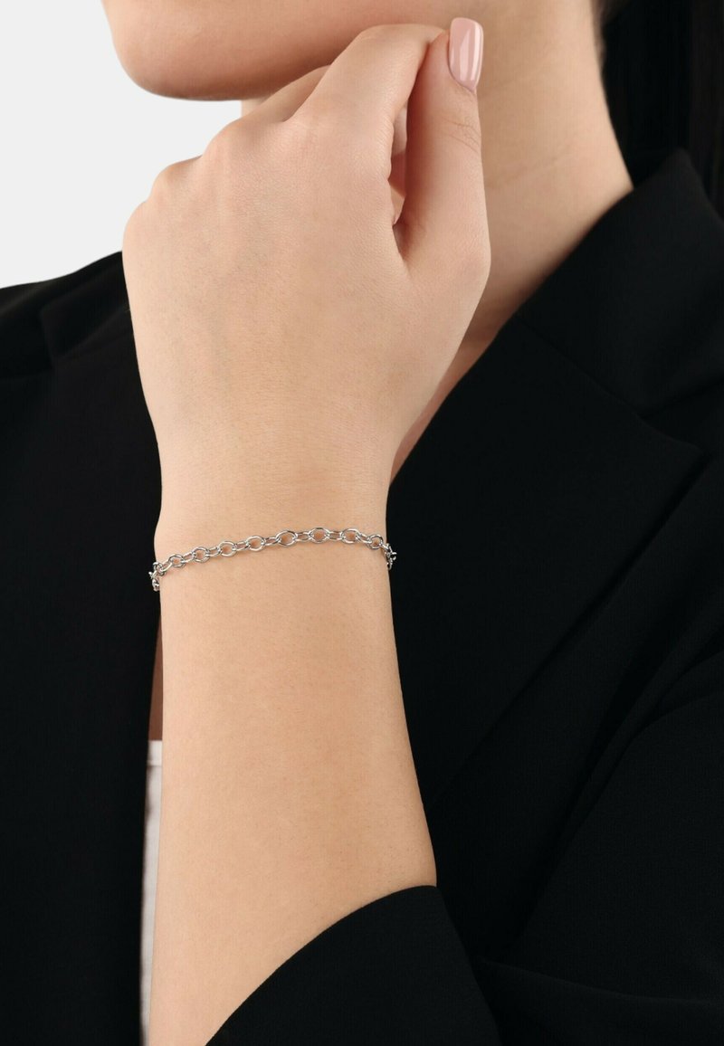 Pulsera de cadena de plata en una muñeca, con eslabones circulares de acabado liso. La mano reposa ligeramente contra una chaqueta negra.