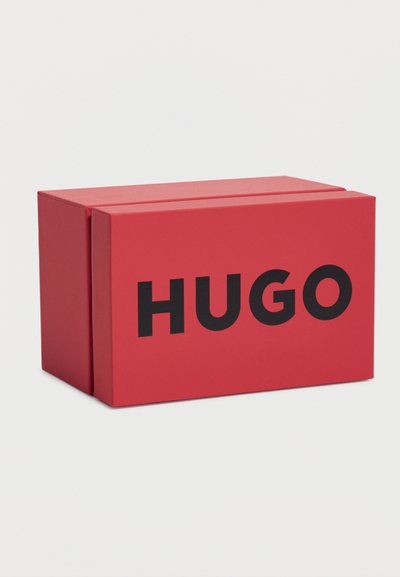 Boîte rectangulaire rouge avec le logo "HUGO" en noir gras sur le devant, posé sur un fond clair uni.