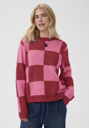 Femme blonde portant un pull à carreaux rose et rouge et un pantalon bleu foncé, une main dans la poche, debout devant un fond uni.