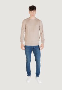Maglione beige a maniche lunghe con collo tondo, abbinato a jeans blu e scarpe da ginnastica bianche. Il maglione presenta un logo discreto sul petto.
