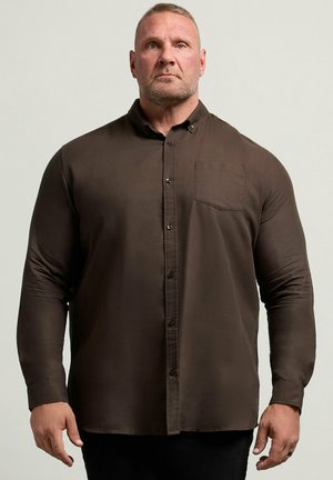Homme aux cheveux blonds courts portant une chemise marron foncé à manches longues et boutonnée, debout devant un fond clair uni.