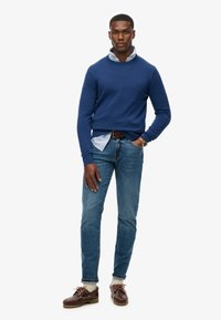 Blå sweater lagdelt over en kravebestrøget stribet skjorte, parret med lyseblå jeans og brune bådsko. Simpelt design, afslappet pasform.