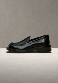 Μαύρο γυαλιστερό δερμάτινο loafer με χαμηλό τακούνι σε ανοιχτό μπεζ επιφάνεια και απλό γκρι φόντο.