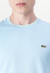 T-shirt en coton bleu clair avec un col rond à côtes, orné de rayures vertes et blanches. Logo de crocodile brodé sur la poitrine gauche.