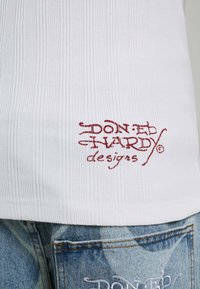 White ribbed shirt with red "Don Ed Hardy designs" logo near hem, paired with faded blue jeans showing partial white embroidered text.