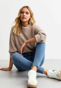 Suéter de punto beige con un cuello redondo amplio, combinado con jeans skinny de mezclilla azul claro con dobladillos deshilachados. Zapatillas blancas con detalles en verde.