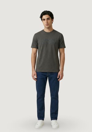 Uomo in piedi che indossa una maglietta Armani Exchange grigio scuro, jeans blu e scarpe da ginnastica bianche su sfondo chiaro uniforme.