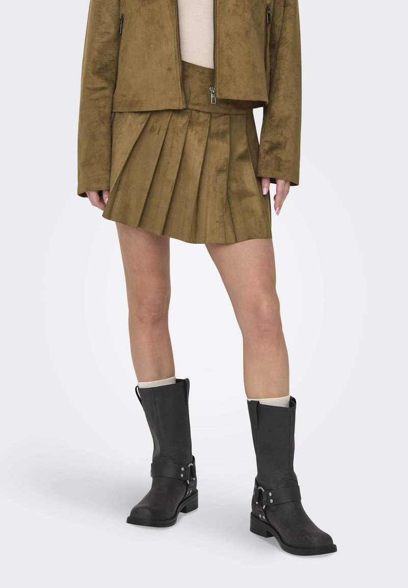 Jupe plissée en daim marron, veste assortie et bottines noires à cheville avec sangles et quincaillerie en métal. Texture lisse et design ajusté.