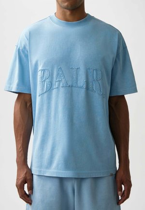 Mann mit hellblauem oversized T-Shirt und passenden Shorts mit großem, strukturiertem "BALR."-Schriftzug auf der Brust.