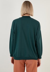 Felpa verde scuro con collo rotondo, spalle scese e polsini a costine. Tessuto liscio con vestibilità comoda. Abbinata a pantaloni marrone chiaro.