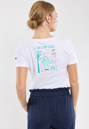 Femme aux cheveux blonds attachés en chignon portant un t-shirt blanc "Le Cap Coz" avec des graphismes colorés sur le thème de la plage et un pantalon bleu marine.