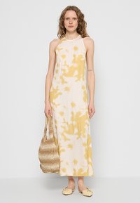 Maxi sukienka z lekkiego materiału o żółto-kremowym abstrakcyjnym wzorze. Charakteryzuje się wysokim dekoltem i bocznym rozcięciem. Dodatki obejmują plecioną torebkę.