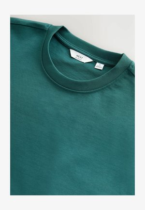Teal crew-neck t-shirt vist på flad, med et mærke der læser "NEXT." Baggrunden er en lys grå farve.