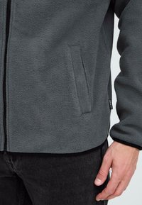 Nahaufnahme einer Person, die eine graue Fleecejacke mit seitlichen Taschen und einem sichtbaren schwarzen Reißverschluss trägt, kombiniert mit schwarzen Hosen.
