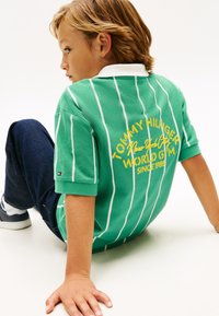 Πράσινο ριγέ πόλο με λευκό γιακά και κίτρινο λογότυπο κειμένου στην πλάτη. Σκούρα μπλε τζιν και λευκά αθλητικά παπούτσια συμπληρώνουν το ντύσιμο.