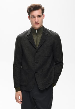 Jeune homme portant un blazer gris foncé sur un pull vert zippé et un pantalon noir, debout avec une main dans la poche.