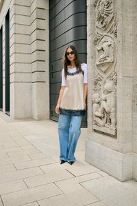 Femme portant des lunettes de soleil, une chemise blanche, une robe beige à dentelle et un jean, se tenant à côté d'une colonne en pierre ornée et de la façade d'un bâtiment.