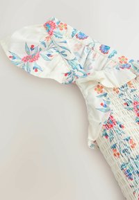 Biely textil s kvetinovým vzorom v modrej, červenej a ružovej farbe, s volánmi na rukáve a detailom nariaseného živôtika.