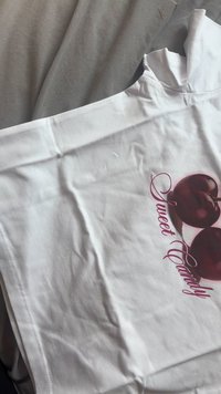 Camiseta de algodón blanca con un gráfico de dos cerezas sobre el texto "Dulce Caramelo" en una escritura rosa. Textura suave, diseño casual.