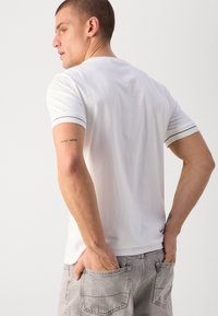T-shirt bianco in cotone con bordi scuri a contrasto sulle maniche, vestibilità comoda e tatuaggio visibile sul braccio. Indossato con jeans di denim grigi.